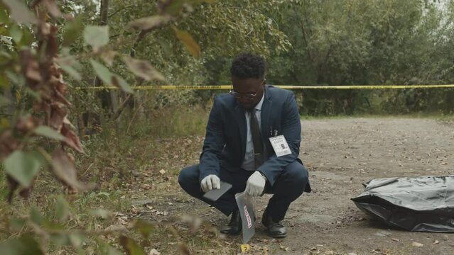 Full length shot of African American male crime investigator in blue suit and gloves collecting evidence beside dead body under black plastic back lying on ground in park