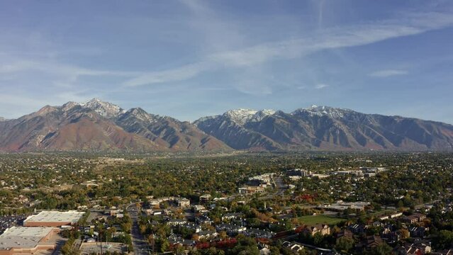 Right trucking tilt down aerial drone wide landscape shot of the stunning snowcapped rocky mountains of Utah with Salt Lake county below full of buildings and colorful trees on a warm sunny fall day