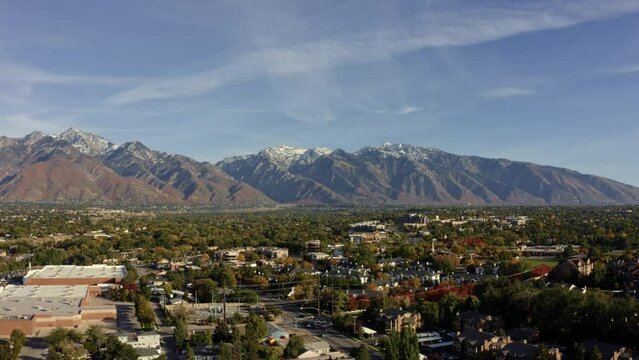 Trucking left aerial drone wide landscape shot of the stunning snowcapped rocky mountains of Utah with Salt Lake county below full of houses and colorful trees on a warm sunny fall day