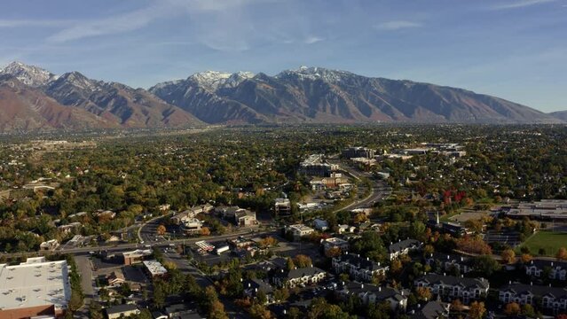 Tilt up aerial drone wide landscape shot of the stunning snowcapped rocky mountains of Utah with Salt Lake county below full of buildings and colorful trees on a warm sunny fall day