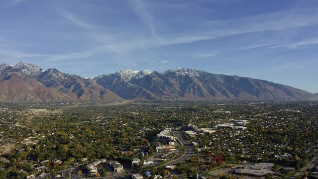 Aerial drone wide landscape shot of the stunning snowcapped rocky mountains of Utah with Salt Lake county below full of buildings and colorful trees on a warm sunny fall day