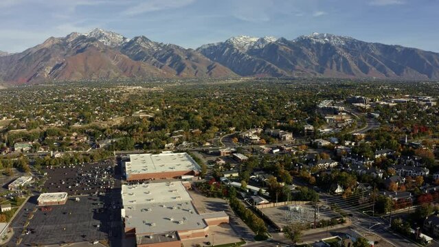 Tilt up right trucking aerial drone wide landscape shot of the stunning snowcapped rocky mountains of Utah with Salt Lake county below full of buildings and colorful trees on a warm sunny fall day