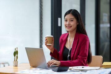 Confident Asian businesswoman holding a paper coffee cup drinking coffee while taking a break at her desk working on corporate financial business documents on a laptop computer.