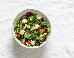 Arugula, cherry tomatoes, cucumbers, mozzarella cheese salad with lemon-mustard dressing on a light background, top view