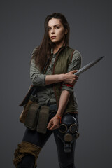 Middle Eastern descent in post-apocalyptic survival gear, brandishing a dagger on a gray backdrop, showcasing her determination to survive