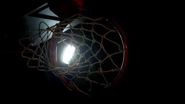 A basketball backboard with a hoop illuminated only by a flickering faulty lamp on the ceiling of a large, dark sports arena. Seamless looping vertical video