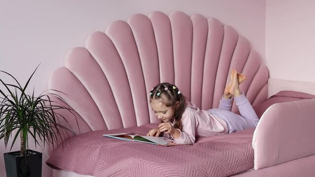 A little girl is lying on the bed looking at the book in slow motion. What to choose a book or a smartphone. The concept of childhood and leisure.