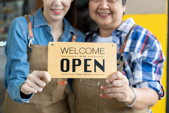 Closeup holding a welcome sign of Young businesswoman, owner of an Asian coffee shop and senior mother.  blue shirt Wear a brown apron. smile happily In front of the counter of a small coffee shop.