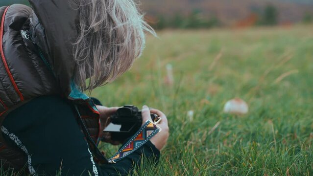 Close up shot of a woman with grey hair from behind lying on the grass and taking photos of a mushroom in nature during a cold windy day in autumn. Shot with Sony FX3 in 4K slow motion