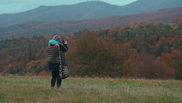 Older female Photographer with grey hair taking pictures with her camera in nature surrounded by autumn coloured trees and grass during a cold windy day in slow motion. Shot with Sony FX3 in 4K.