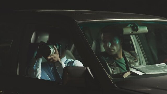 Waist up of two multiethnic male cops stalking and tracing down criminal in car at night. Black male officer taking photos on professional camera and showing them to colleague at driver seat