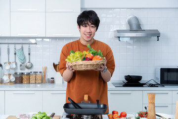 Happy portrait of asian young man holding a basket of vegetables of standing a cheerful preparing food and enjoy cook cooking with vegetables, while standing on a kitchen Condo life or home
