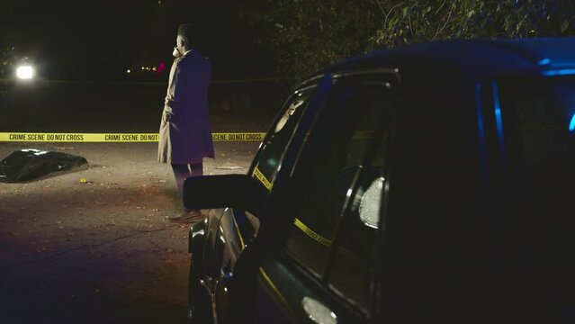 Back view full shot of African American male detective in long camel trench coat crossing yellow barrier tape and examining murder scene at night. Corpse in black plastic bag lying on ground