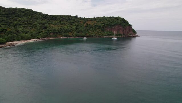 Frontal drone flight from sea to Punta de Mita's rocky shores, with boats and a lush green mountain contrasted by blue sea.