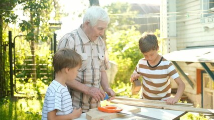 An elderly gray-haired man teaches boys carpentry outdoor.