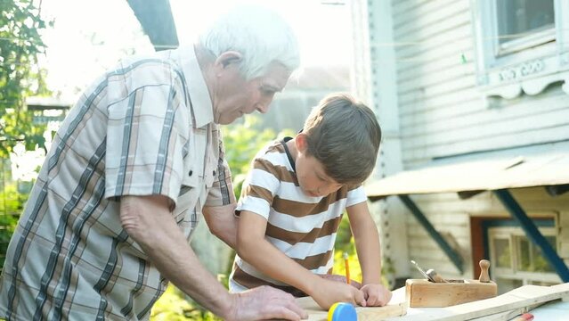 An elderly gray-haired man teaches a boy carpentry outdoors. Grandfather teaches his grandson to carpenter, measure a wooden board with a ruler and draw a line with a pencil.