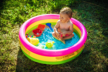 A cute little baby boy is playing with toys while bathing in a little colorful wading pool on meadow garden field outdoor. Summer time. Joy and happiness. Love and family