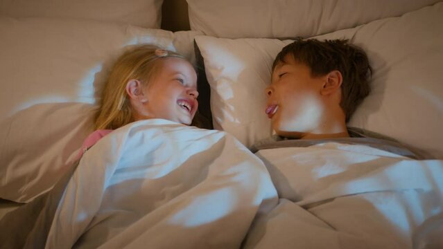 Funny Caucasian older brother make little sister laugh show tongue grimacing in bed before bedtime happy laughing children kids boy girl playing together having fun sibling affection relationship