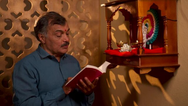 elderly senior man reading hymns from religious book in front of god near mandapam at home at morning - concept of religious custom, idol worshipping and spirituality