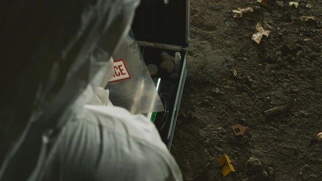 From above shot of anonymous forensic specialist in protective coveralls collecting evidence from ground in plastic bag and putting it into kit at murder scene at night