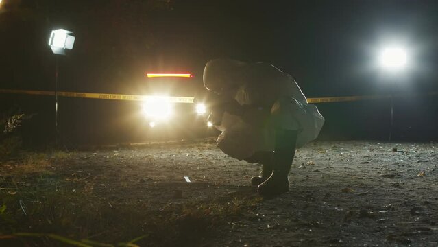 Anonymous forensic expert wearing protective coveralls, respirator mask and gloves taking photographs of crime scene on camera while working at night