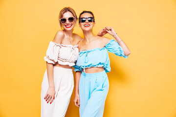 Two young beautiful smiling brunette hipster female in trendy summer clothes. Sexy carefree women posing near yellow wall in studio. Positive models having fun. Cheerful and happy. In sunglasses