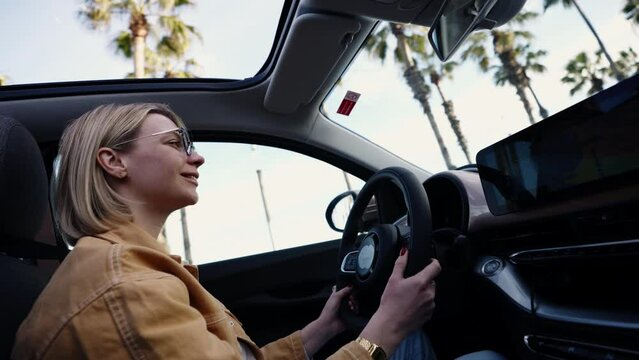 Blonde woman driving car with palm trees visible through open sunroof, clear sky, concept of freedom and leisure in a modern urban setting