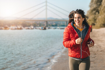 Happy young woman jogging during the chilly autumn day on the sandy beach while wearing casual...