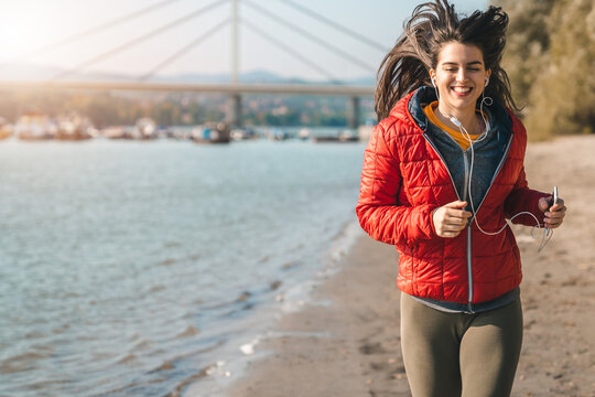 Attractive smiling female fitness instructor running at the seaside on a sunny cold day in autumn.
