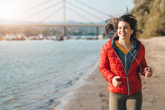 Young smiling sporty fitness woman wearing a red sport jacket and sweatshirt running on seaside while holding phone and listening to music.