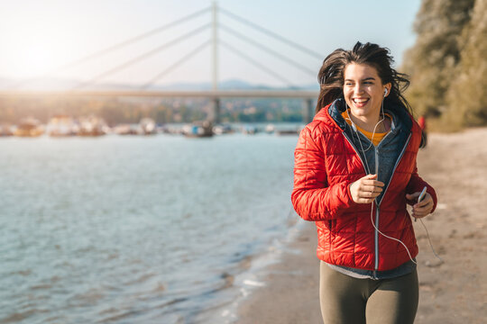 Happy young woman jogging during the chilly autumn day on the sandy beach while wearing casual sportswear and headphones.