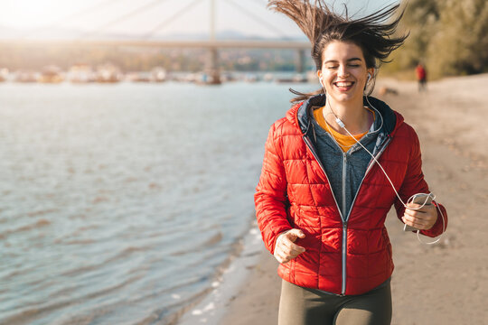 Young smiling sporty fitness woman wearing a red sport jacket and sweatshirt running on seaside while holding phone and listening to music.