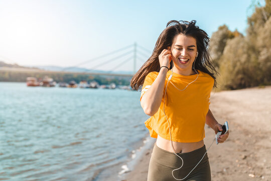 Cheerful woman in yellow t-shirt running on the beach and enjoying healthy lifestyle and music outdoors during the sunny spring morning.