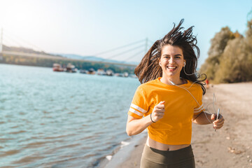 Young smiling woman in yellow t-shirt with long hair looking at camera jogging on seaside while...