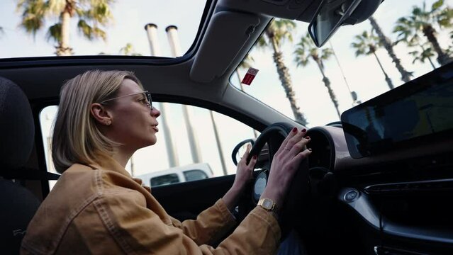 Blonde woman clapping hands in car, expressing excitement or approval, sunroof open to blue sky and palm trees, concept of celebration and joy while driving