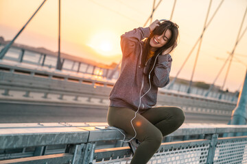 Beautiful young sportswoman smiling while listening to music on a bridge in the morning. Fit woman enjoying her favorite song.