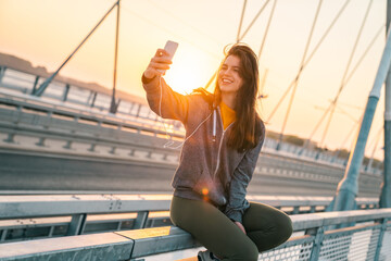 Beautiful young female taking a selfie while sitting on the bridge at sunrise. Attractive woman enjoying early morning exercise outdoors.