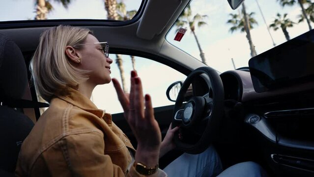Blonde woman gesturing with hand while driving and singing song, open sunroof with palm trees in view, concept of relaxed communication and enjoyable drives