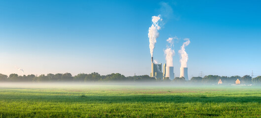 Coal Power Plant in green meadow with morning fog smoking and steaming against blue sky