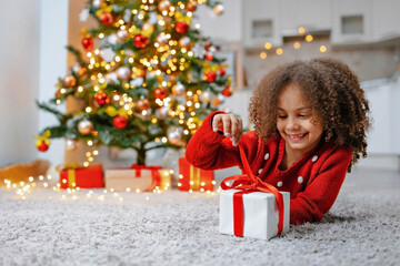 Fototapeta na wymiar A Black girl in a red sweater lies on the floor and unties a bow on a gift box near a decorated Christmas tree.