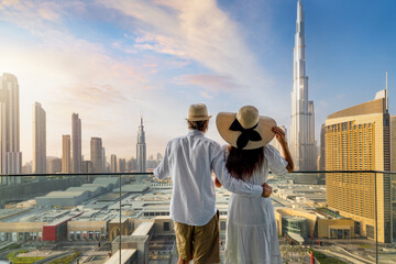 A elegant tourist couple on vacation time stands on a balcony and enjoys the sunrise view of the Dubai city skyline, UAE - obrazy, fototapety, plakaty