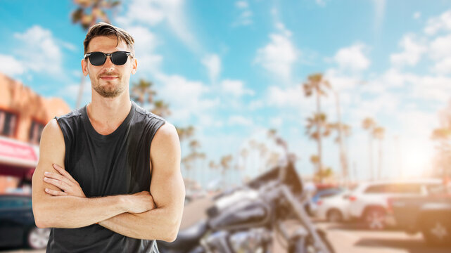 Portrait of American young handsome man chopper biker rider  in sleeveless shirt with sunglasses summer coast beach background standing smiling.