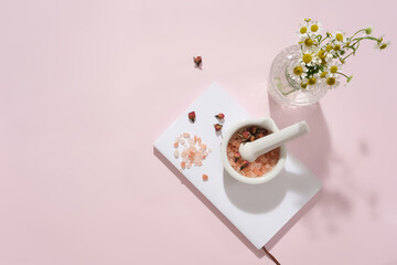 A bouquet of daisies is placed in a glass jar, pink salt is placed in a white ceramic mortar on a pastel pink background. Pink salt contains many minerals that help clean the skin.