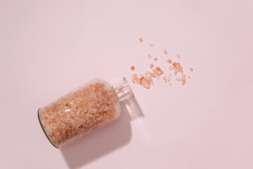 Pink salt grains are poured into a pastel pink background from a transparent glass bottle. Himalayan pink salt is a type of rock salt originating from the Punjab region of Pakistan.