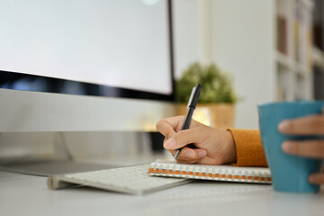 Side view of woman sitting in front of computer monitor and writing notes in personal daily planner