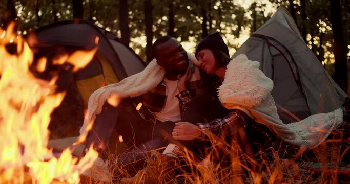 Happy couple: a girl with a bob hairstyle examines the face of a guy with black skin, touches him and leans on his shoulder, they hug under a white blanket, sit near a bright fire against the backdrop