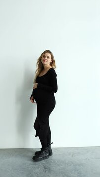 a pregnant girl in a black dress on a white background enters the frame, poses and exits the frame. holds his hands on his stomach and walks. vertical video