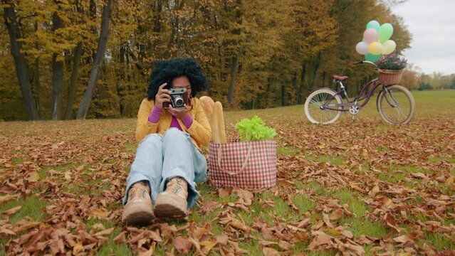 Picturesque park with yellow trees and fallen leaves. Smiling woman sitting on grass with camera, paper bag with products next to her, bicycle behind. High quality 4k footage