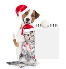 Happy Jack russell terrier puppy and funny cute kitten wearing santa hats standing together. Dog shows empty notepad. Cat holds candy cane. isolated on white background