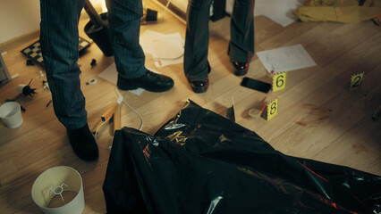 Two detectives are standing near a corpse in a bag at the crime scene, studying the evidence